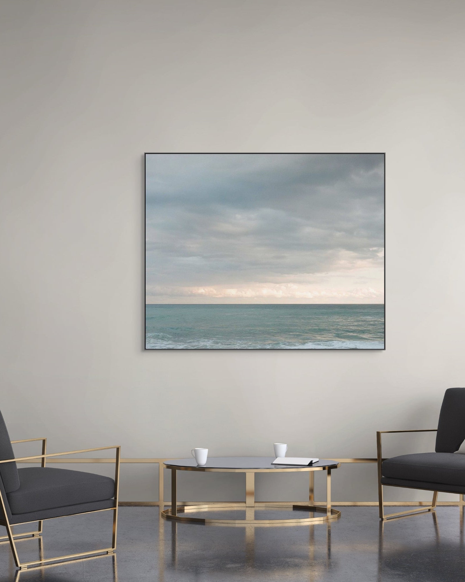 Seascape photograph with a thin black frame displayed in a modern lounge area with gold-accented chairs, a round glass coffee table, and soft grey walls.