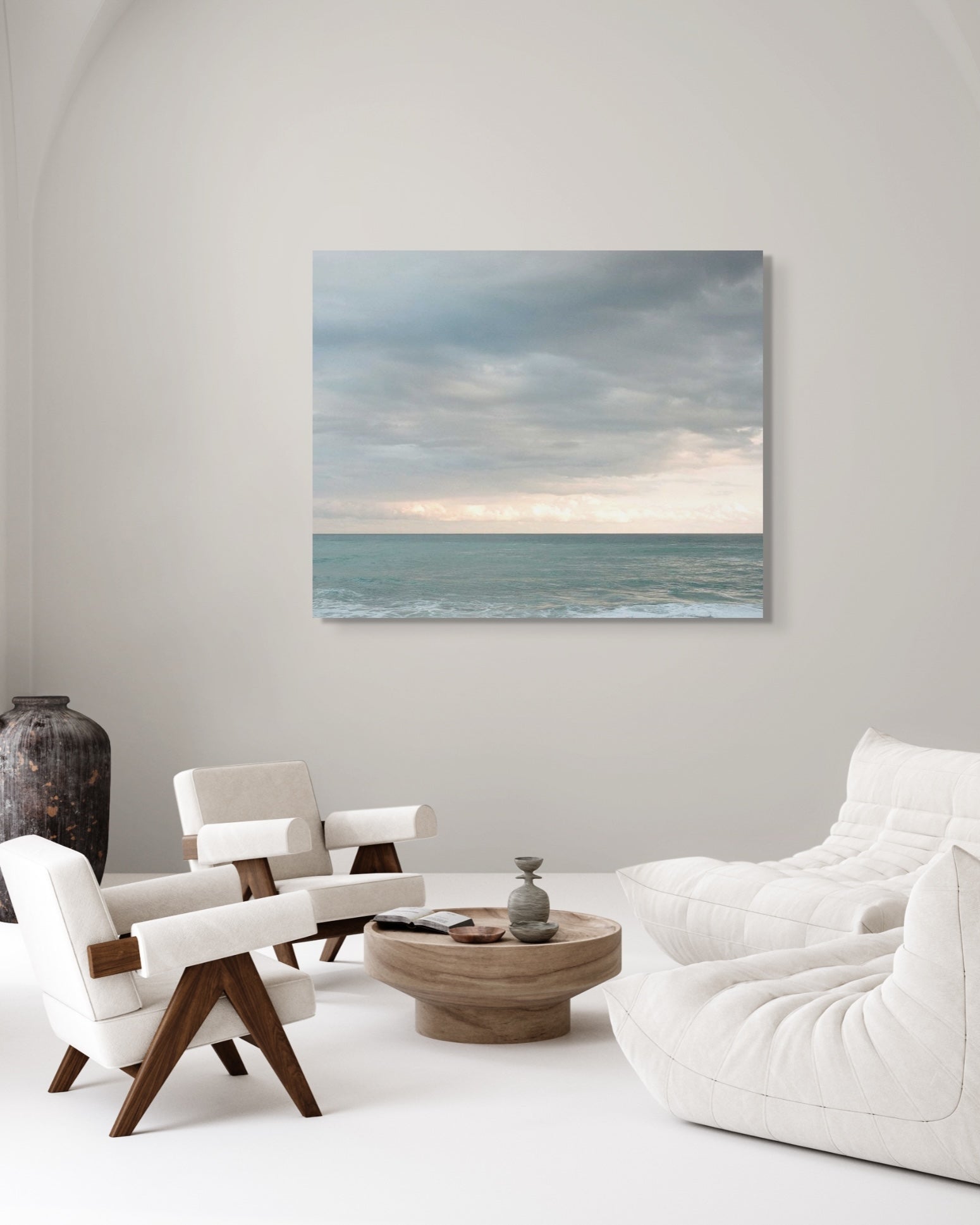 Framed seascape photograph hanging above a modern cream-toned living room with soft lounge chairs and a round wood coffee table.