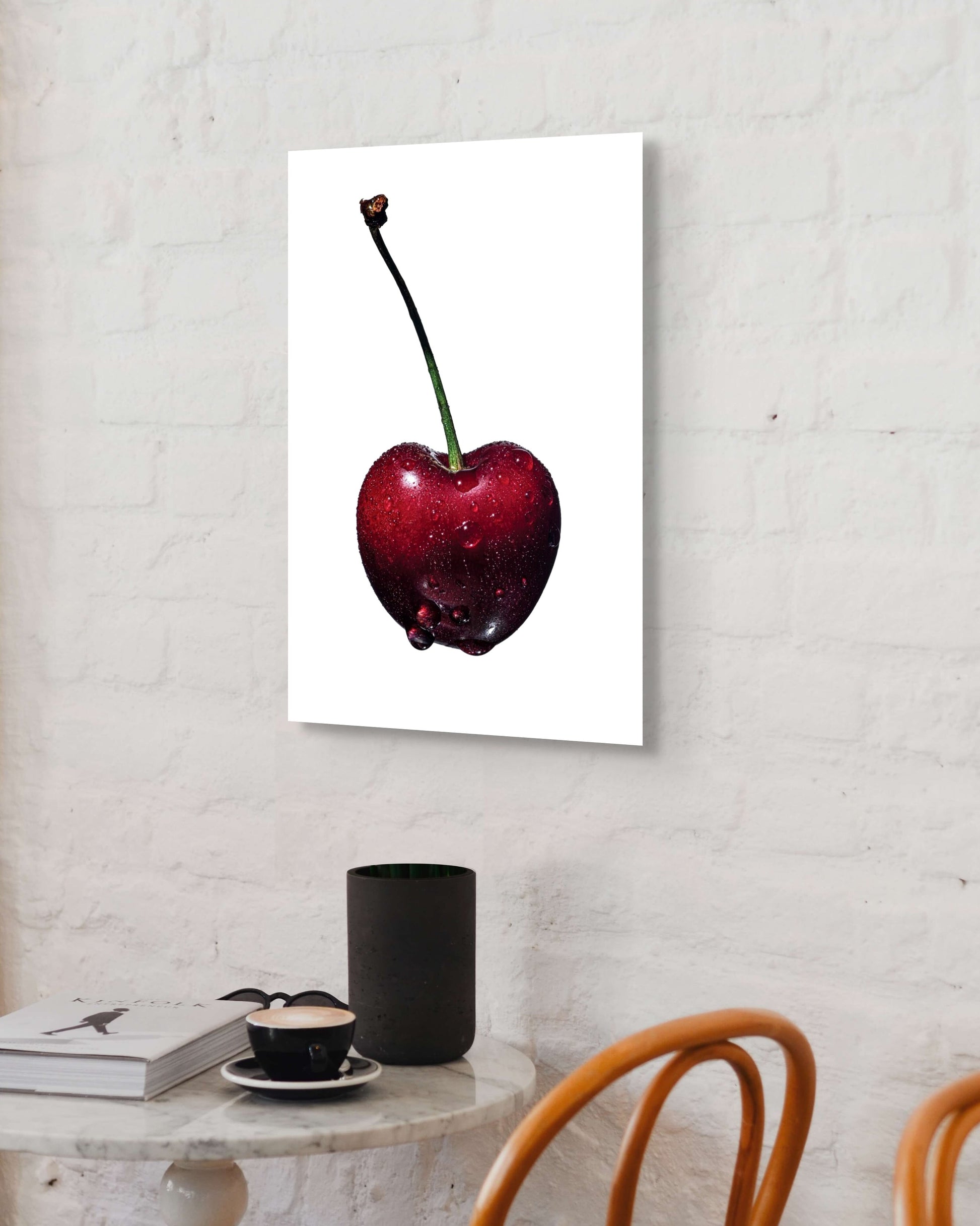 Metal print of a red cherry on a white wall above a table with a cup and books.