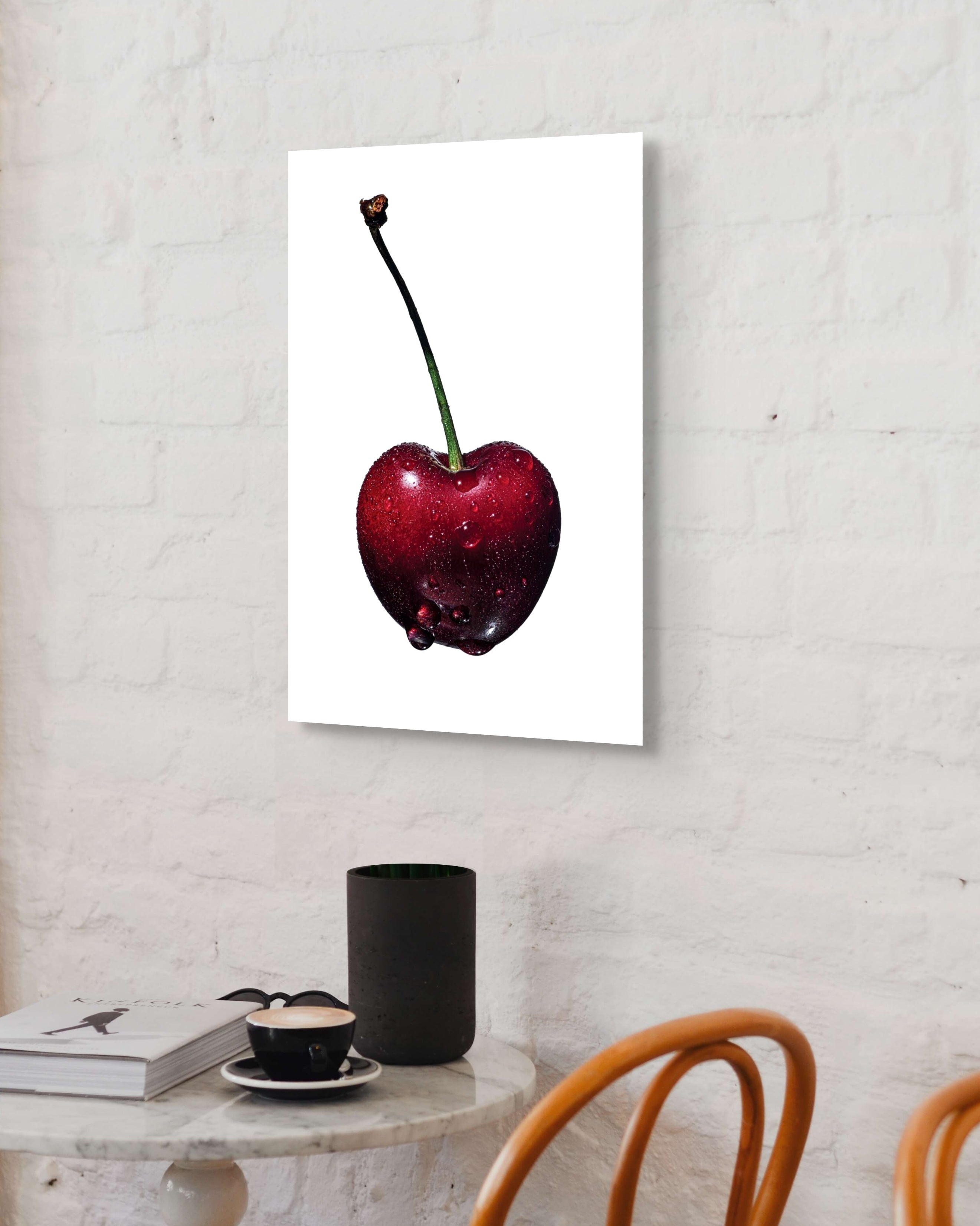 Metal print of a red cherry on a white wall above a table with a cup and books.