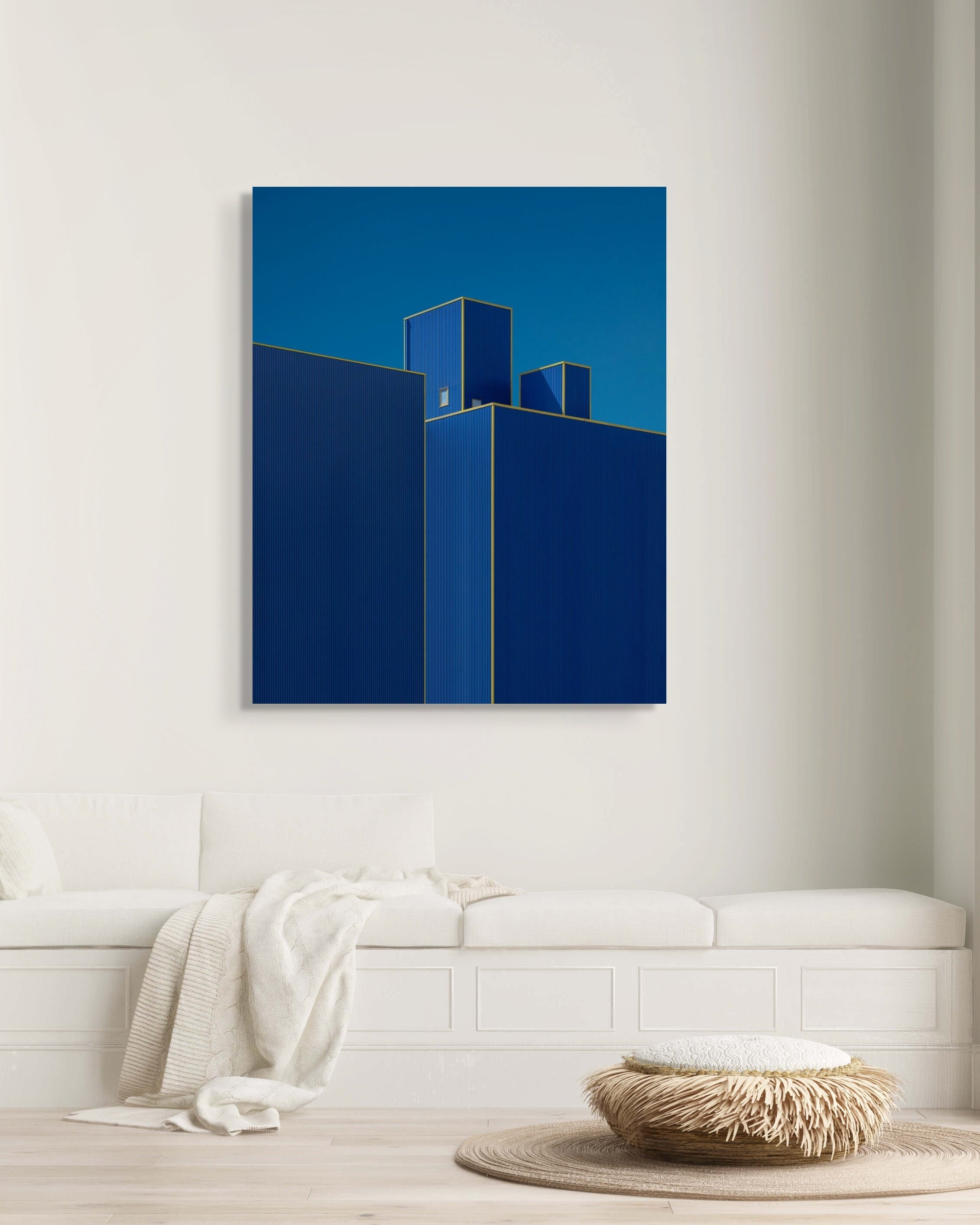 A large metal print of “Blueberry II” by Mark Forbes, featuring deep blue geometric building forms with gold outlines, displayed above a white built-in bench in a minimalist living room.