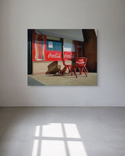 Framed picture of a store exterior with Coca-Cola bottles and chairs on a white wall.
