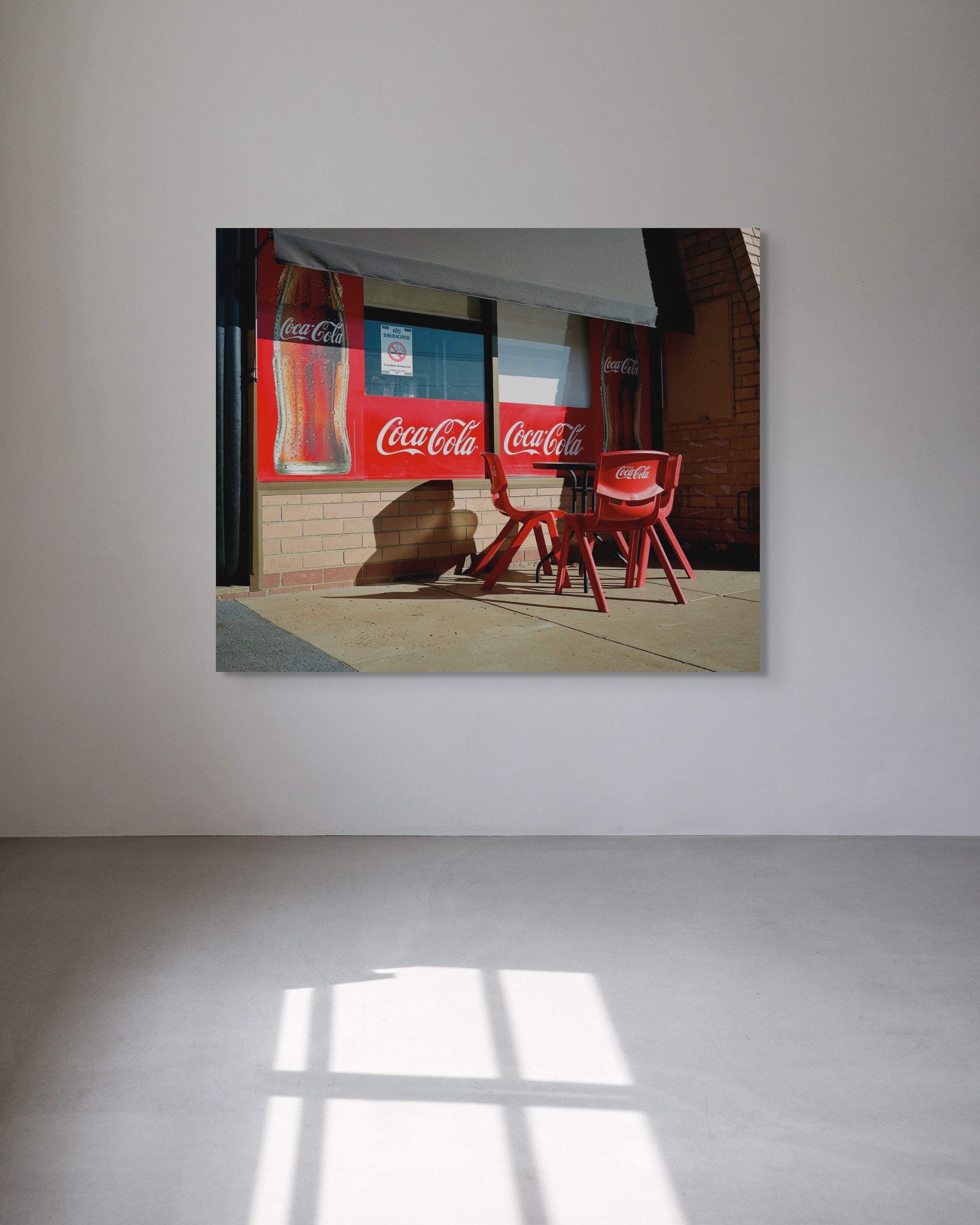 Framed picture of a store exterior with Coca-Cola bottles and chairs on a white wall.