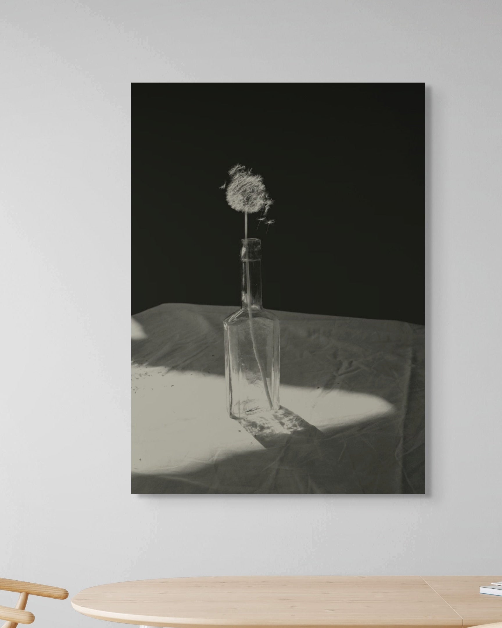 Framed black and white artwork of a bottle with a dandelion on a gray wall above a wooden table.