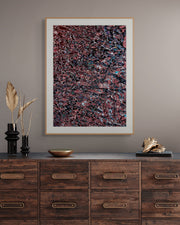 Framed abstract photograph with a white mat and gold frame displayed above a rustic wood console, featuring deep reds, blacks, and subtle blue accents in an intricate, fluid pattern.