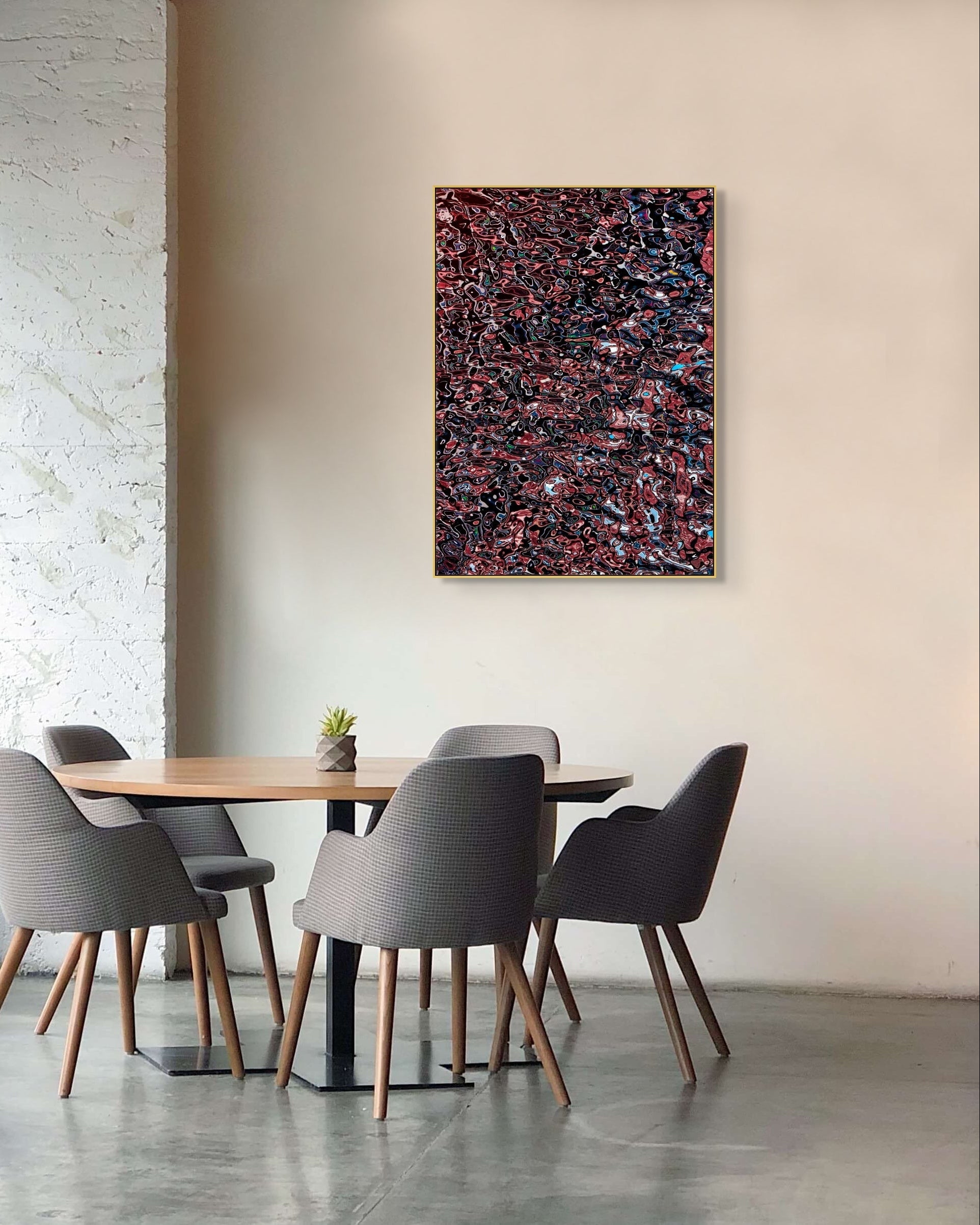 Abstract photographic print in a thin gold frame hanging on a light wall in a modern café-style seating area, showing layered red, black, and blue textures with a reflective, fluid appearance.