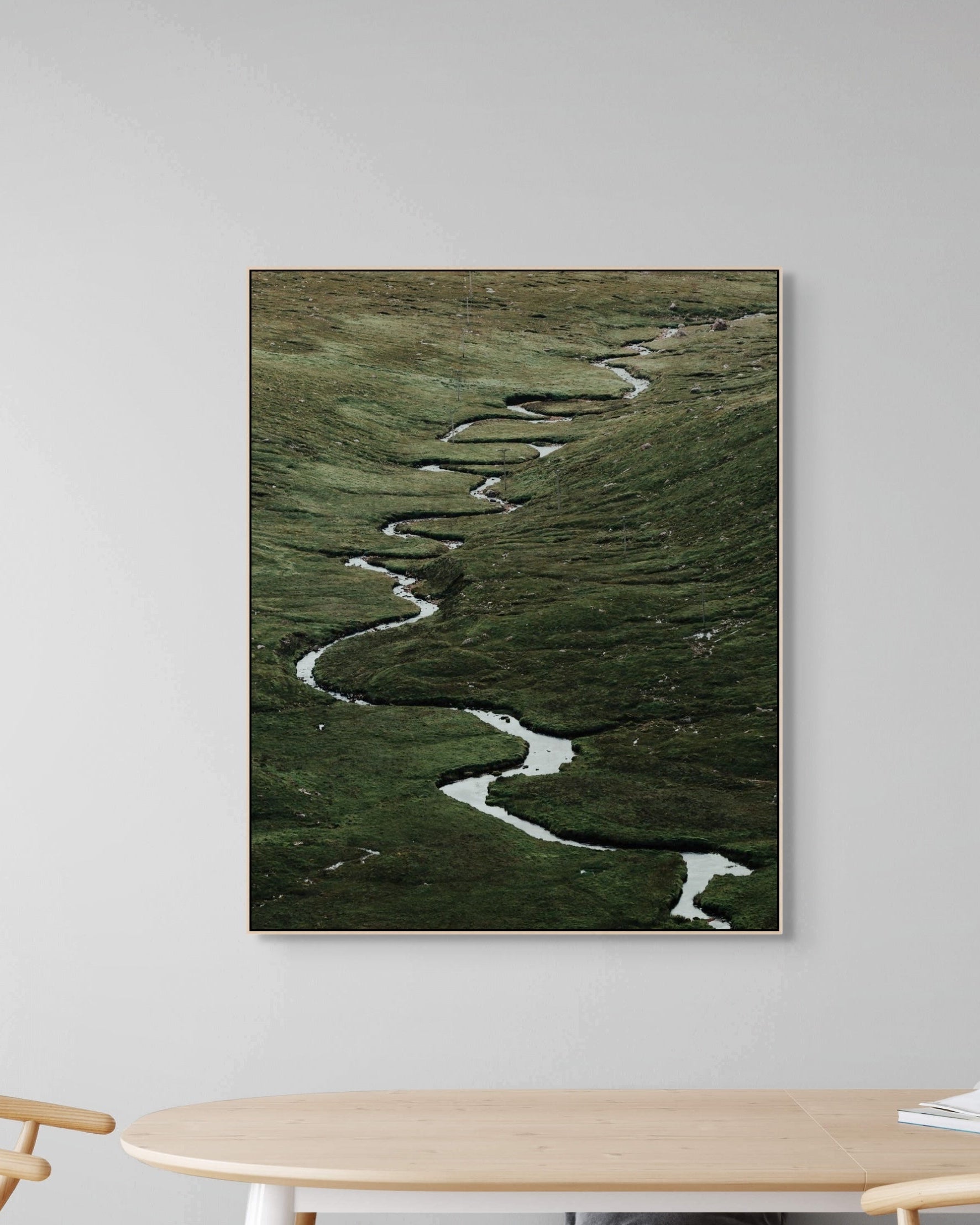 Framed artwork of a winding river on a grassy landscape, displayed on a wall above a table.