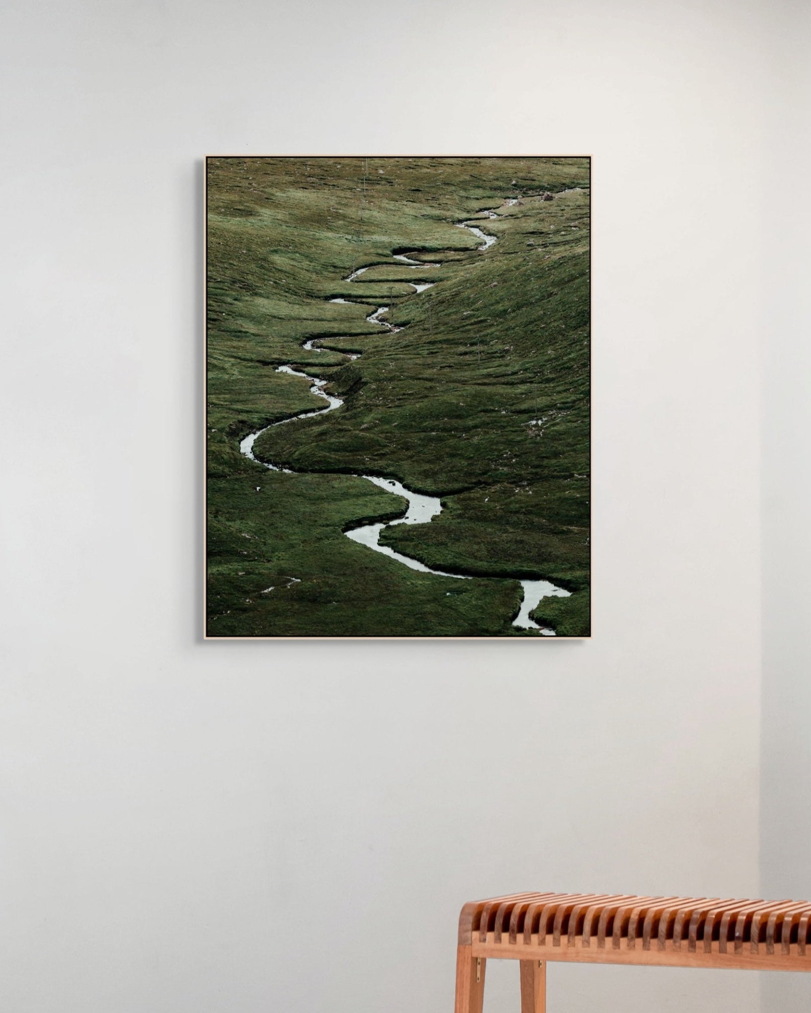Framed artwork of a winding river on a grassy landscape, displayed on a wall above a wooden bench.