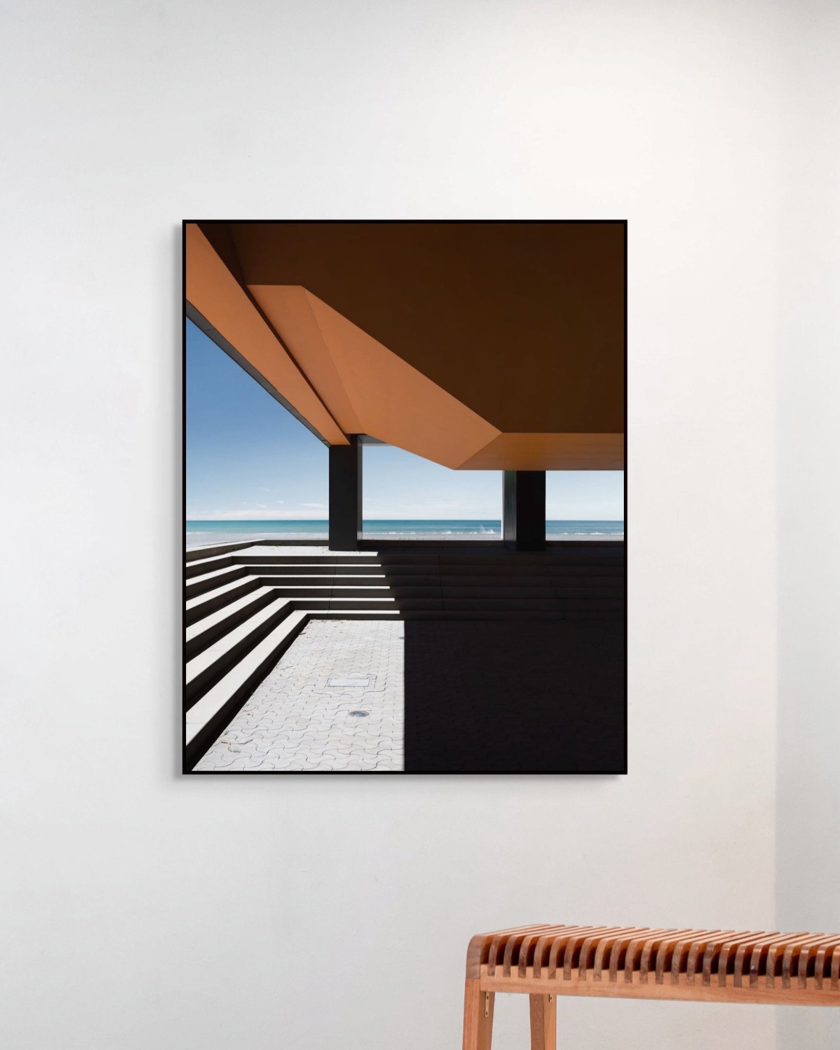 Framed artwork of a modern architectural design on a white wall with a wooden bench below.