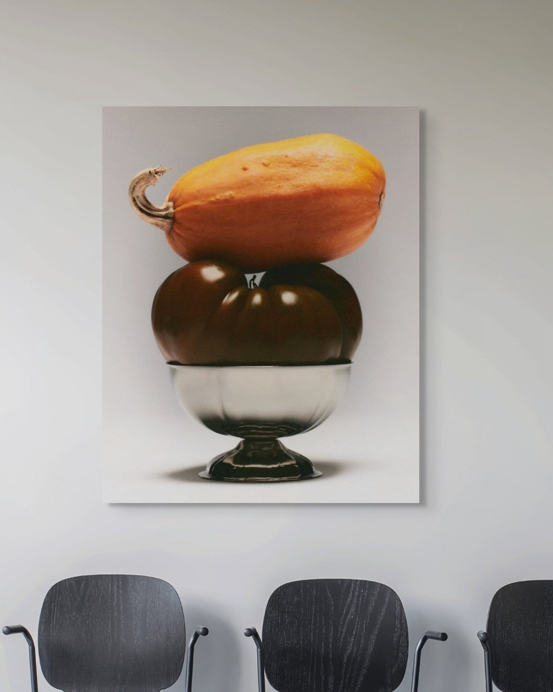 Framed artwork of two stacked pumpkins on a white wall above three black chairs.
