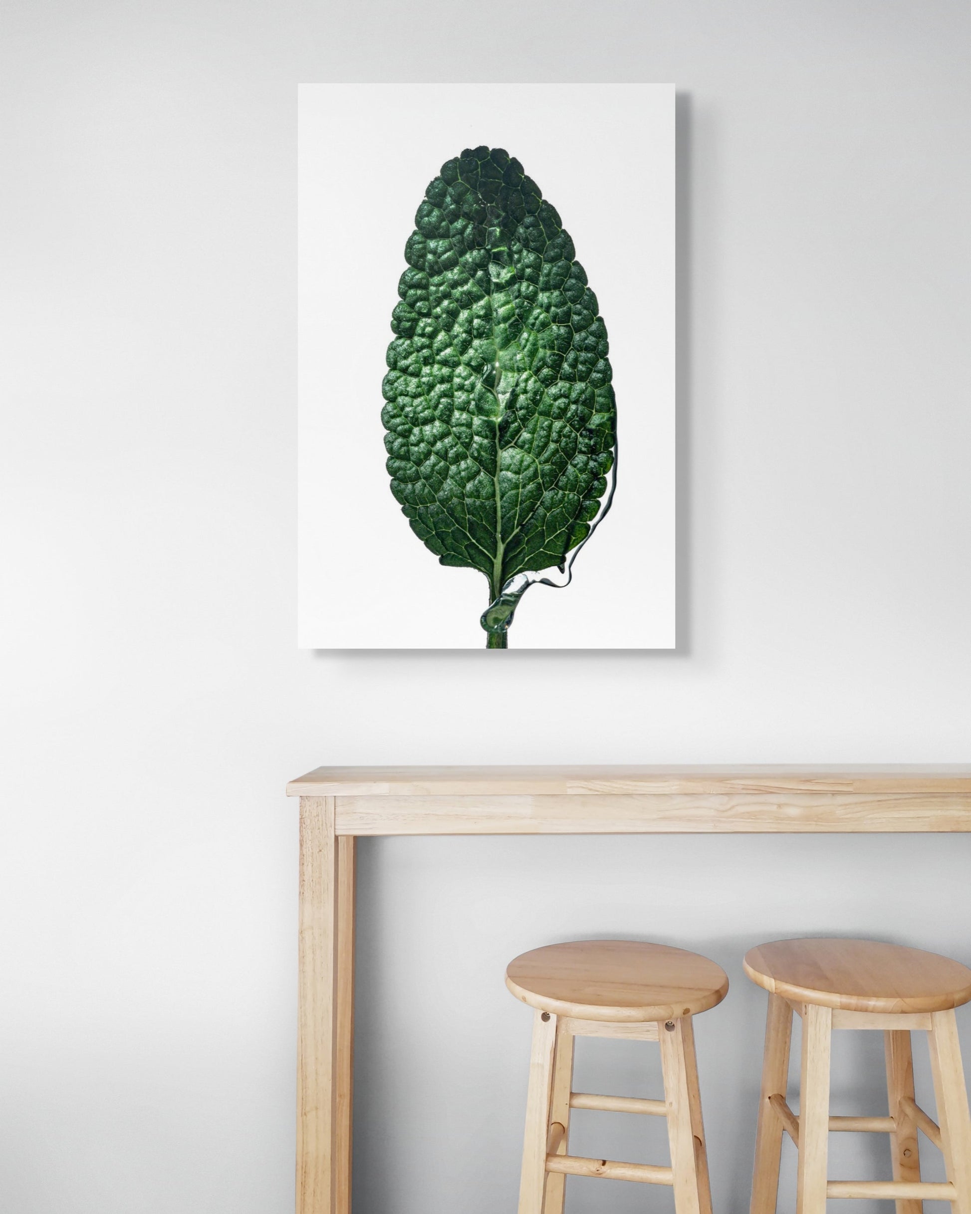 Sage Leaf by Sivan Roshianu – Framed and ready to hang limited edition fine art photograph of a green leaf on a white wall above a wooden table with stools.