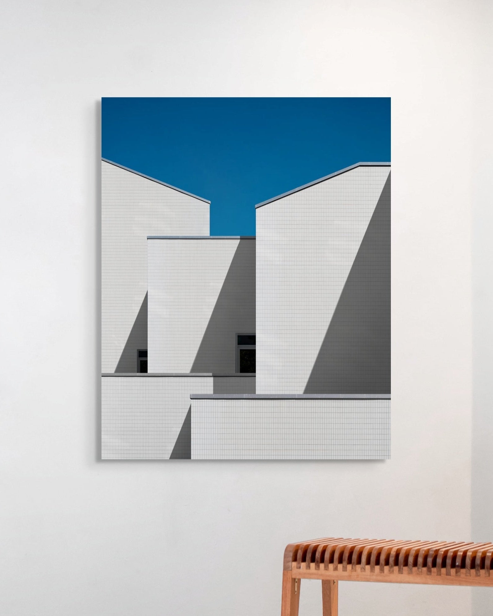 Abstract art piece with geometric shapes on a blue background, displayed on a white wall with a wooden bench below.