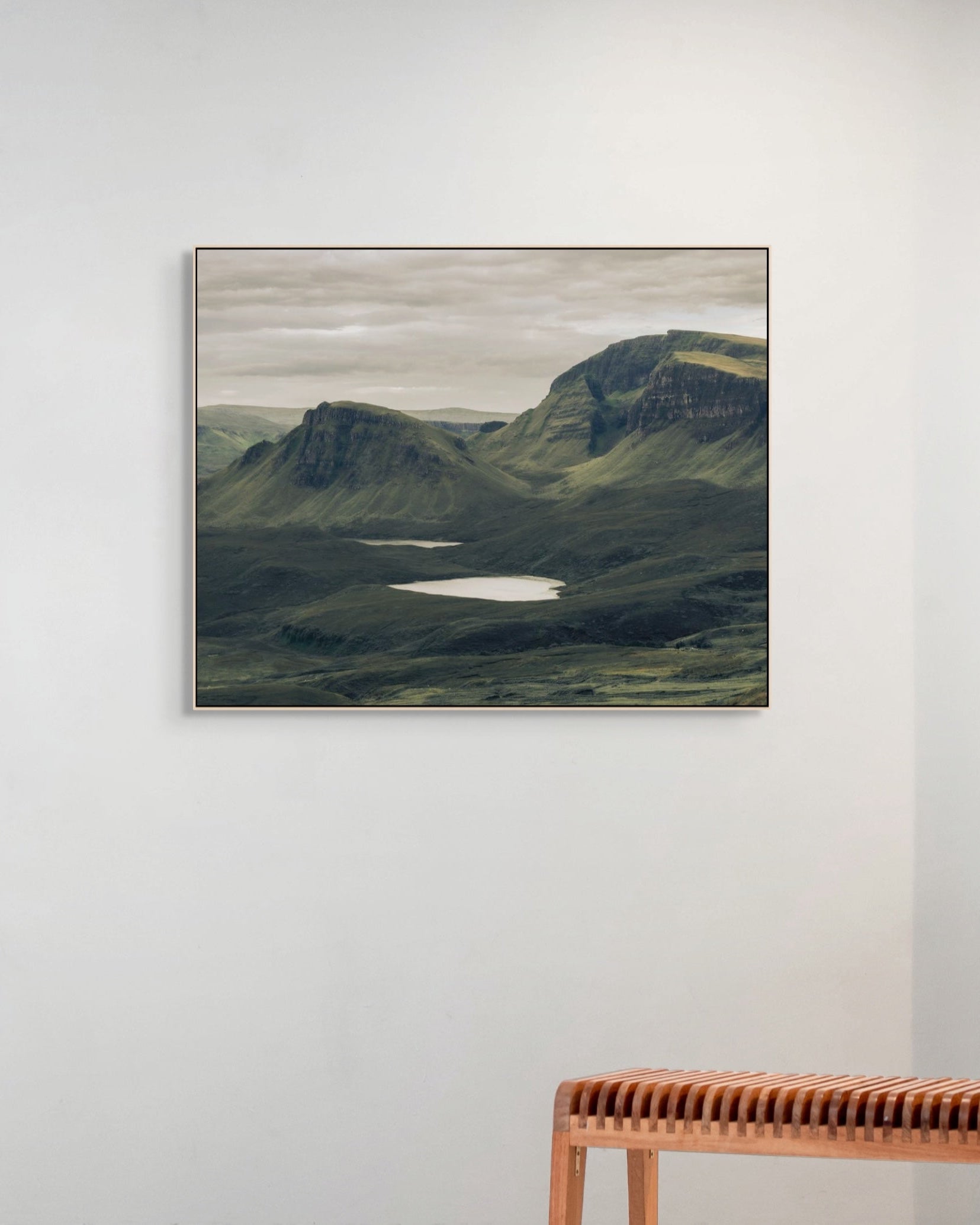 Framed landscape print of a mountainous scene with a lake on a white wall above a wooden bench.